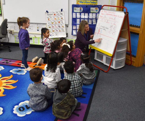 A teacher writes on an easel dry erase board while young students crowd around sitting on the floor watching.