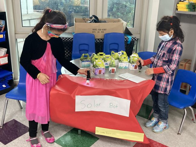 Two girls in face masks to a hands on activity on opposite sides of a school table.
