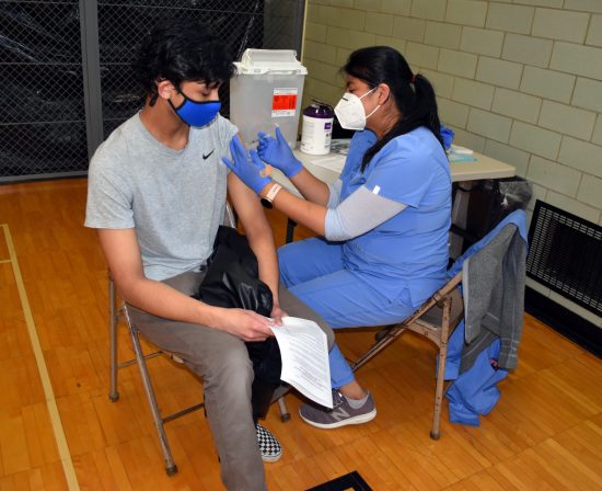 A student watches as a nurse gives him a vaccination shot in the shoulder during a COVID vaccination clinic at Edsel Ford High School in May 2021.