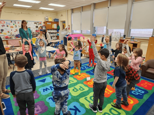 A group of young students stands and stretches their arms over their heads inside a preschool classroom.