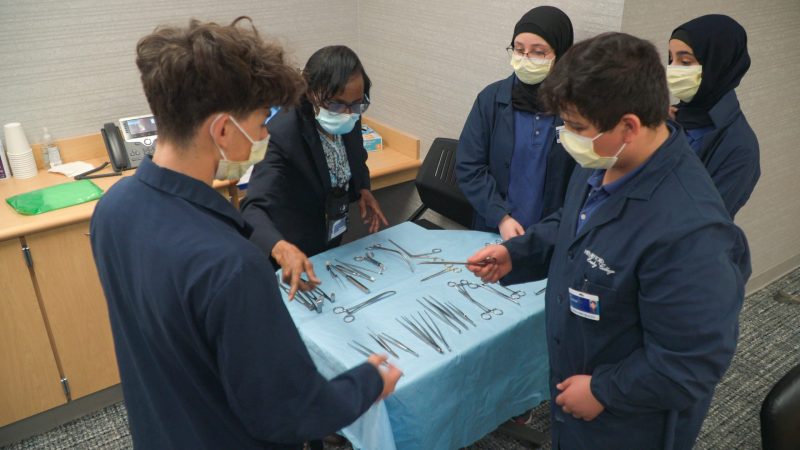 Four Henry Ford Early College students and a woman stand around a surgical tray as students learn more about the instruments and how to handle them.