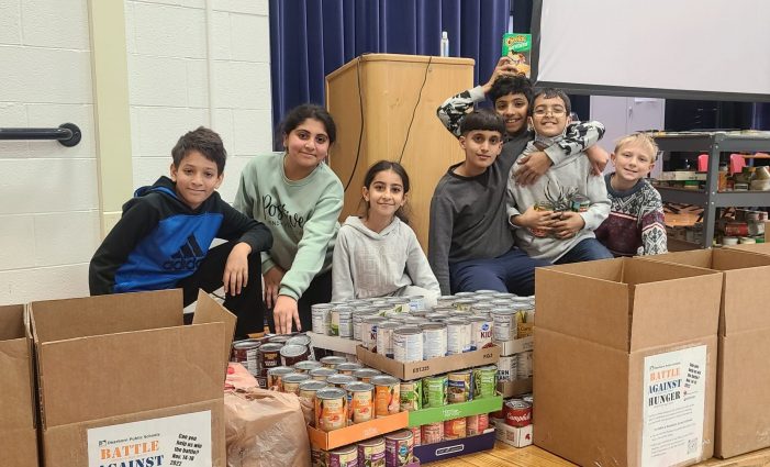 Six students sit on a school stage surrounded by box and trays of donated food.