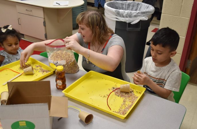 A woman helps two young children with a bird feeder craft.