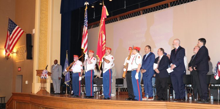 A group of adults stands on stage while the colors are presented during a Memorial Day Ceremony at Fordson High.