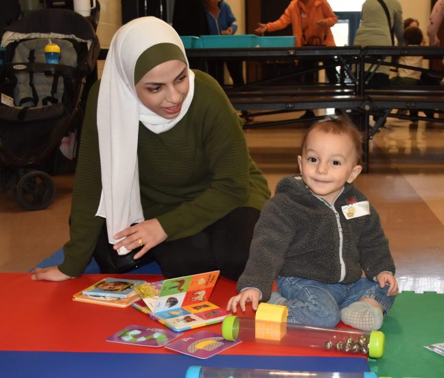 A woman sits on a mat next to a baby surrounded by toys and board books.