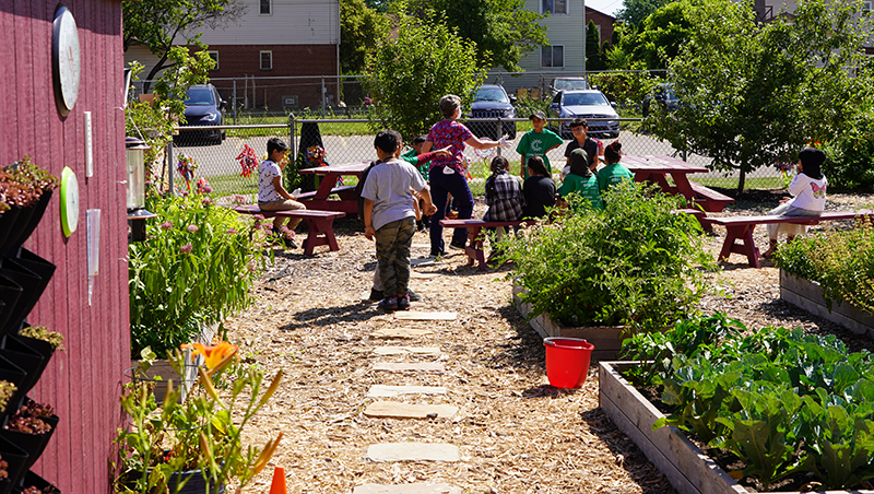 A teacher instructs students sitting on benches in a school garden