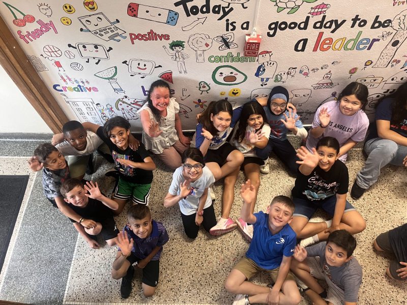 More than a dozen students sit in a school hallway by a mural they are working on that says "Today is a good day to be a leader."